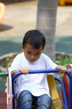 Küçük bir çocuk oyun parkındaki ekipmanların üzerinde oturur, renkli bir açık hava ortamında masumiyet ve oyunculuğu yansıtan bir anı ve neşeyi yakalar..
