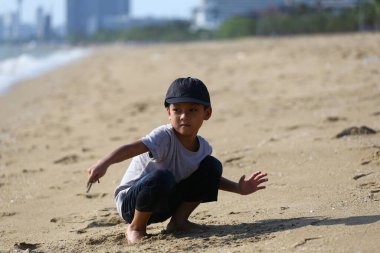 Neşeli genç bir çocuk kumsalda güneşli bir günün tadını çıkarıyor, kumsalda neşeli bir ifadeyle oynuyor, etrafı okyanus ve kıyı şeridiyle çevrili. Aile ve yaz temaları için mükemmel.