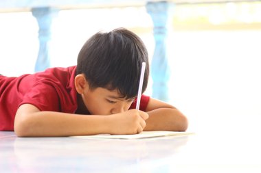 Yerde yatan genç bir çocuk, derinden yazmaya odaklanmış, konsantrasyonunu ve yaratıcılığını doğal ışıkla dolu açık hava ortamında sergiliyor..