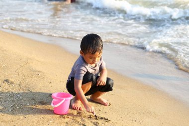 Güzel bir sahilde kumla oynayan neşeli bir çocuk, pembe bir kova kullanarak okyanus kenarında unutulmaz bir deneyim yaratıyor..