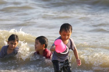 Üç çocuk kumsalda güneşli bir günün tadını çıkarıyorlar, yumuşak dalgalarda oynuyorlar. Bir çocuk pembe bir kova tutarken kahkaha havayı dolduruyor, neşeli anılar yaratıyor..