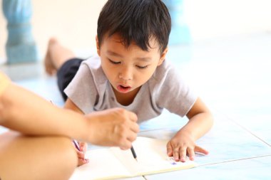 Genç bir çocuk yerde yatarken bir not defterine kalemle çizim yapmaya derinden odaklanır. Bir yetişkinin eli, kapalı alanda yaratıcı keşifleri destekler..