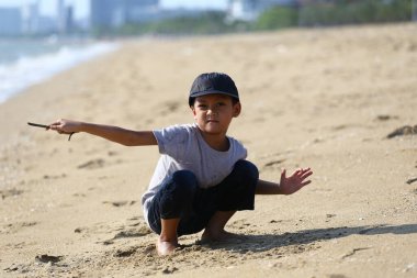 Genç bir çocuk kumsalda neşeyle oynar, kumlara çömelirken sopayı oyuncak olarak kullanır, parlak güneşli bir günde çocukluğun kaygısız ruhunu somutlaştırır..