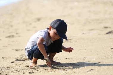 Genç bir çocuk kumsalda güneşli bir günün tadını çıkarıyor. Oynuyor ve yumuşak kumu keşfediyor. Bu görüntü, huzurlu bir sahil ortamında çocukluğun neşesini ve masumiyetini yakalar..