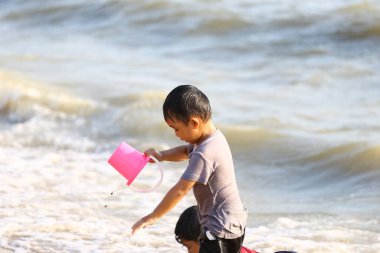 Neşeli bir çocuk sahilde canlı pembe bir kovayla oynar, su sıçratır ve dalgalar yavaşça kıyıya vururken neşeli anlar yaratır..