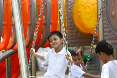 Parlak renkli bir çocuk parkında salıncaklarda eğlenen neşeli bir sahne. Güneşin altında neşe ve beraberlik anları sergiliyorlar..