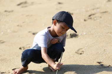 Genç bir çocuk kumsalda güneşli bir günün tadını çıkarıyor, kumla etkileşime girerken derin bir oyun oynuyor. Bu görüntü çocukluk sevincinin ve doğadaki keşiflerin özünü yakalar..