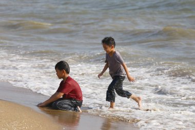 İki neşeli çocuk sahilde oynuyor, bir çocuk sahilde koşarken diğeri kumsalda oturuyor, mükemmel bir yaz günü eğlencesini ve kahkahalarını yakalıyor..