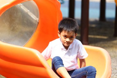 Genç bir çocuk parlak bir turuncu kaydırağın üzerinde oturur, güneşli bir günün sıcaklığının tadını çıkararak, çocukluk sevincinin ve tasasız maceranın özünü yakalar..