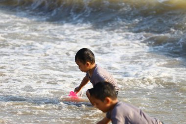 İki neşeli çocuk sahilde sığ sularda oynar, güneşin tadını çıkarırlar ve yaz tatillerinde neşeli anılar yaratarak hafif dalgalarda yüzerler..