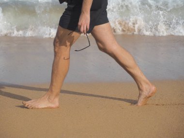 Man walking on the beach, wet feet and sunglasses in hand.