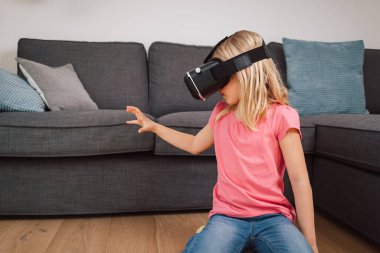 Evde sanal gerçeklik deneyimi sırasında VR kulaklıklı genç bir kız havaya dokunuyor. Artırılmış gerçekliği keşfeden duygusal çocuk.