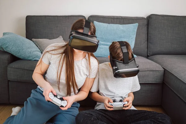 Mother and son enjoy entertainment video gaming together, using VR goggles and controllers while sitting on a sofa in the living room