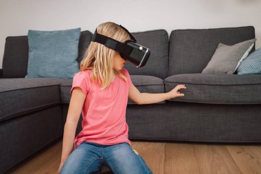 Evde sanal gerçeklik deneyimi sırasında VR kulaklıklı genç bir kız havaya dokunuyor. Artırılmış gerçekliği keşfeden duygusal çocuk.