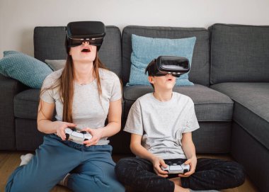 Kafkasyalı kız ve küçük kardeşi evde eğleniyor, sanal gerçeklik kulaklıkları ve kontrol cihazları kullanarak bilgisayar oyunu oynuyorlar.