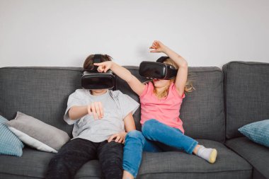Beyaz bir oğlan ve VR kulaklık takan küçük bir kız oturma odasında film izliyor ya da oyun oynuyorlar. Sanal gerçeklik, teknoloji ve çocukluk kavramı.
