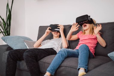 İki çocuk, bir oğlan ve VR kulaklık takan küçük bir kız oturma odasında oturup etkileşimli bir deneyim sayesinde sanal dünyada eğleniyorlar.