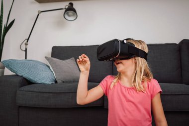 Evde sanal gerçeklik deneyimi sırasında VR kulaklıklı genç bir kız havaya dokunuyor. Artırılmış gerçekliği keşfeden duygusal çocuk.
