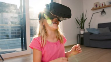 Smiling Caucasian little girl having fun at home, wearing VR goggles enjoying interactive virtual reality experience