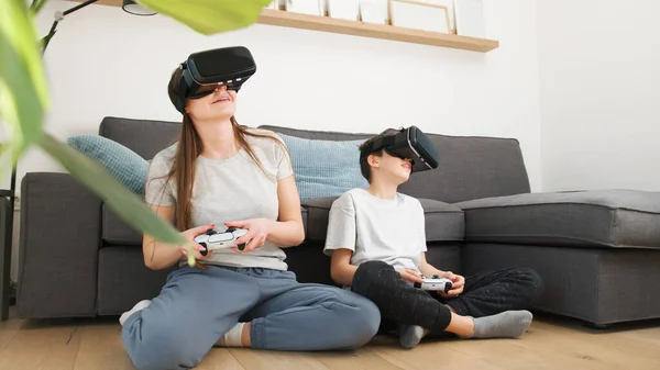 Mother and son enjoy entertainment video gaming together, using VR goggles and controllers while sitting on a sofa in the living room