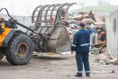 Male worker pointing on wheel loader and the garbage heap at a landfill site, rear view. Waste disposal, consolidation, and transfer concept.