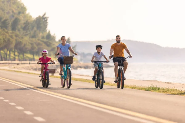 Two kids, with helmets on their heads, and smiling parents riding bikes on a family-friendly cycle route along a sea coastline, front view.