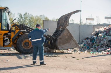 Landfill worker directing skid steer loader on the garbage heap, rear view. Waste disposal, consolidation, and transfer concept.