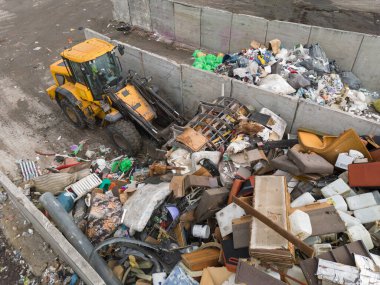 Handling construction waste on the landfill site, skid steer loader scooping and dumping dusty trash, aerial side view.