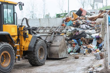 Yellow wheel loader, with lifted scrap grapple, moving along the recycling center area in process handling dumped waste
