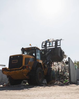 Yellow wheel loader, with lifted scrap grapple, moving along the recycling center area in process handling dumped waste