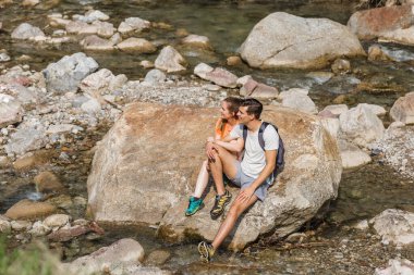 Young couple taking a break from hiking, sitting on the mountain river bank and watching beautiful nature