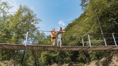 Loving couple, recreational hikers, crossing a suspension bridge over a rocky mountain river bed. Adventure and travel concept.