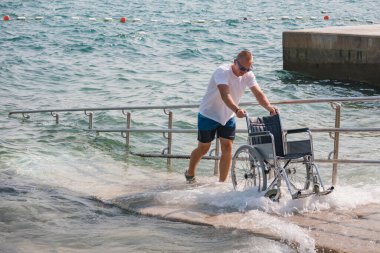 Cankurtaran engelliler için rampası olan erişilebilir plajda tekerlekli sandalyeyi denize itiyor.