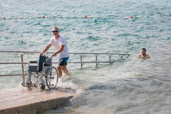 Man with disability on wheelchair at accessible beach goes swimming.