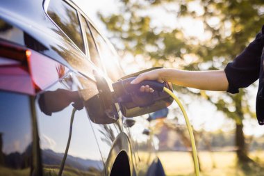Siyah elektrikli bir arabanın şarj aletini takan genç bir kadın, yenilenebilir enerji kavramı.
