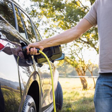 Genç adam siyah elektrikli bir arabanın şarj aletini takıyor, yenilenebilir enerji kavramı.