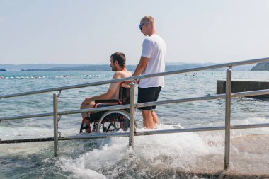 Plajda engelli bir adam, tekerlekli sandalyede yüzmeye gider. Erişilebilir bir rampadan yardım alarak..