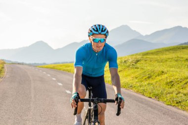 Mavi formalı ve teçhizatlı bir sporcu kaldırımlı yolda profesyonel bir yarış bisikletine biniyor.