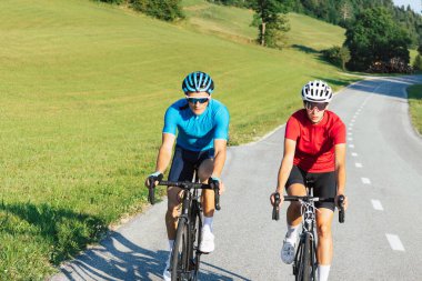 Bir kadın ve bir erkeğin ön görüntüsü, spor giyimli profesyonel yarış bisikletçileri, asfalt bir yolda güzel yeşil doğada ilerliyorlar..
