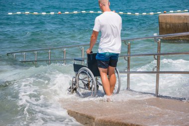 Cankurtaran engelliler için rampası olan erişilebilir plajda tekerlekli sandalyeyi denize itiyor.