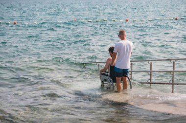 Plajda engelli bir adam, tekerlekli sandalyede yüzmeye gider. Erişilebilir bir rampadan yardım alarak..