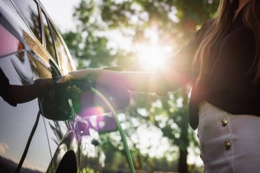 Kadın el, güneş ışınlarıyla aydınlatılan elektrikli bir arabaya kablo şarj aleti yerleştiriyor yapraklı ağacın dallarını kırıyor.