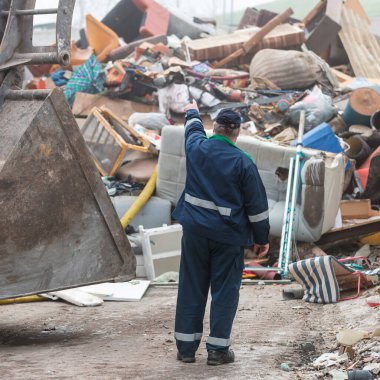 Worker at a recycling center standing near a bucket loader, looking and pointing at the waste heap, rear view.
