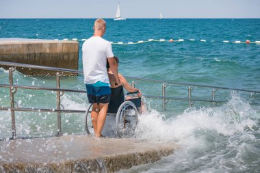 Plajda engelli bir kadın, tekerlekli sandalyede yüzmeye gider. Erişilebilir bir rampadan yardım alır..