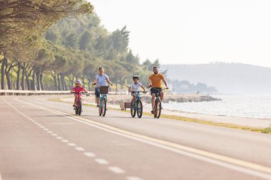 Two kids, with helmets on their heads, and smiling parents riding bikes on a family-friendly cycle route along a sea coastline, front view.