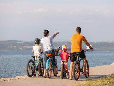 Genç bir aile, iki çocuklu ebeveynler, deniz kenarında duran bisikletlerle, annenin işaret ettiği yöne bakıyorlar.