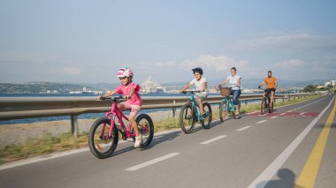 Dört kişilik bir aile, oğlu ve küçük kızı olan ebeveynler deniz manzaralı bir sahil yolunda bisiklet sürerken rahatlar.
