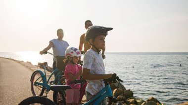 Caucasian family with two children standing on bicycles and enjoying the sunset on a seashore, rear view.