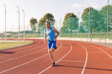 Genç bir adam, ciddi ve odaklanmış bir yüzü olan profesyonel bir atlet spor sahasının yakınındaki açık hava atletizm pistinde koşuyor.