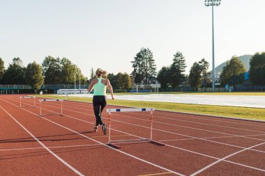 Genç şişman kadın güneşli bir yaz gününde atletizm pistinde koşuyor ve engellerin üzerinden atlıyor.